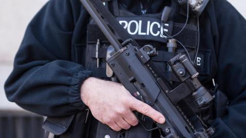 An armed police officer's mid section. He is dressed in black with a bullet-proof police vest on and is holding a large gun