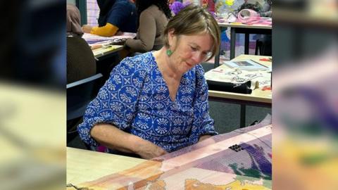 Dr Lynn Setterington wearing a blue dress with white circular print on it, sitting at a table working on an embroidery piece. There are embroidery materials in the background.