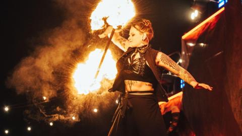 A female fire performer standing on stage as she holds up a baton lit with fire either end in one of her hands. She has tattoos and is wearing a black outfit. The photo has been taken at night.