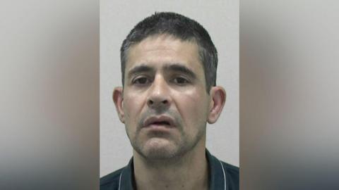 Mugshot of Andreas Michael. He has a wonky mouth, faint stubble and short dark hair tinged with grey.