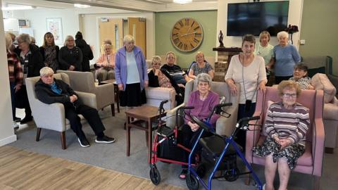 A group of elderly men and women sit in a large open-plan living area...