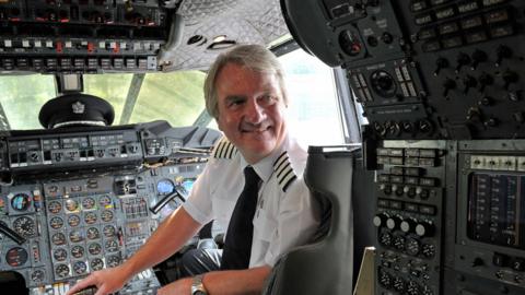 A pilot, Mike Bannister, sat in a plane cockpit and looking behind him. He is looking beyond the camera.