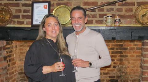 Sam Davies has long brown hair and is wearing a black blouse, standing next to David Wheeler, who is also holding a flute glass. They are both smiling inside, with a bricked fireplace behind them.