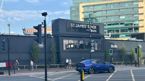 St James' Stack. It is a made up of a number of repurposed shipping containers painted black. A car drives past while a number of people are walking outside.