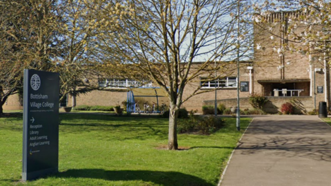 A google maps image of the front of Bottisham Village College. There is a pathway leading to the front doors of the building which is made from a light brown coloured brick. Next to the pathway is a grassy area with a sign for the collage and trees behind it. There is also a blue bike shed at the end of the grassy area, just in front of the school building. 