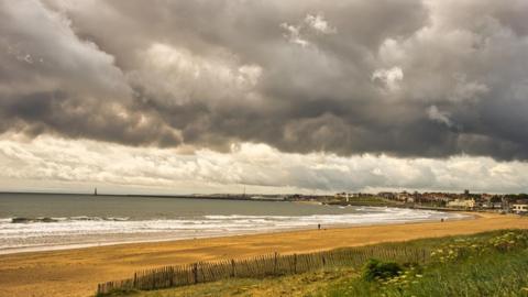 Grey, cloudy skies over the sea with surf, sand and grass in the foreground