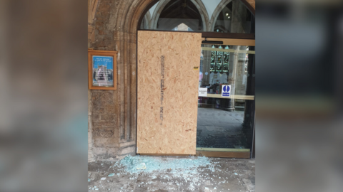 An arched stone entrance to a church which has been partially covered up by a wooden board. Beneath the board are many fragments of light blue shattered glass. To the right of the board is a second automatic door which is still intact.