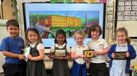 A group of six small children, two boys and four girls, stand holding picture frames with artwork in them. Behind them is a large image of another artwork.  They all wear school uniforms in blue, grey and white colours.  The artwork behind them depicts a large mill building with chimney tower. The pictures they are holding also depict flowers and nature.