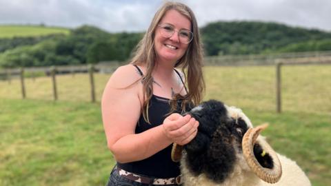 Woman with long brown hair wearing glasses and smiling at the camera with her  large ram with curly horns 