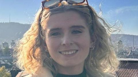 Síofra Harkin has long blonde curly hair and is wearing brown and black sunglasses, nestled on her forehead. She is wearing a green top and a beige jacket. 