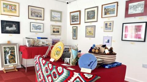 A table with painted plates and a red patterned table cloth, with paintings of scenery in the background.