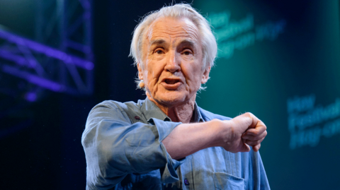 Larry Lamb in a blue shirt gesticulating towards the audience while on stage at Hay