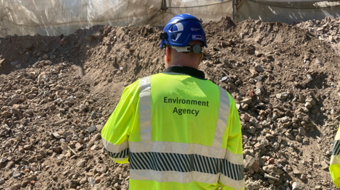 A worker in an Environment Agency fluorescent jacket and a helmet inspects a mound of rubble and soil.
