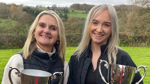 Two women are standing in a field holding silver trophies. Both have blonde hair, the one on the left has a jacket with a dark centre section and cream sleeves while to our right, the other woman has a black jacket over a black top. They stand in a  field with trees behind them and more trees in the distance.