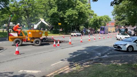A picture of the roadworks near Hyde Park Corner in Leeds, there is a digger in the middle of the road with a workman sitting on it, there are cones on the road, with cars and buses and trees in the background.  