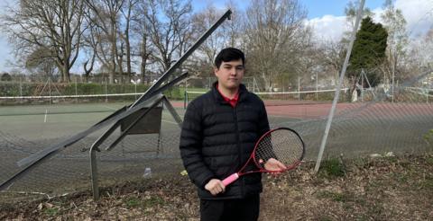 A man with dark hair and wearing a black jacket and trousers, red T-shirt and holding a tennis racket stands in front of the mangled fence with tennis courts behind it, 