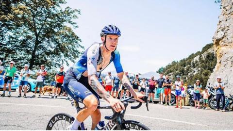 Jake Stewart in athletic clothing riding a bike on a road lined with spectators in the sun