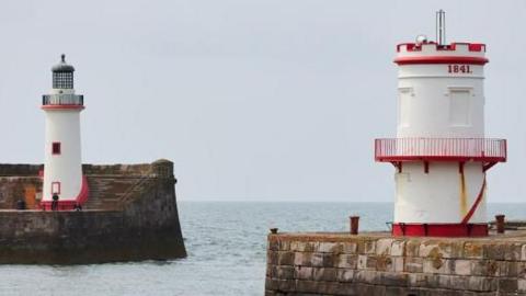 A view of the lighthouses on the ends of two piers at Whitehaven Harbour. The lighthouses are white and painted red around the railings.