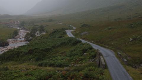 A large green landscape with a river on the left of the image and a road running  on the right hand side. The road disappears into the distance and it's a misty day.