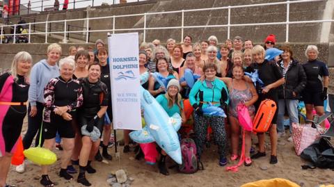 The Hornsea Dolphins - a large group of women in wetsuits standing on a beach. Some hold bright orange floatation devices and inflatable animals.