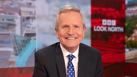 Presenter Peter Levy sat in the Look North studios with a suit on and smiling at the camera. He has grey hair and is wearing a black suit jacket, white shirt and blue tie with white spots. 