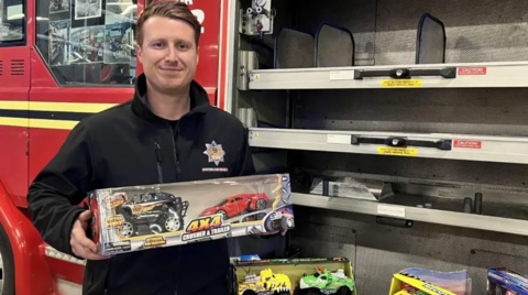 A man wearing a black jumper stands next to a fire engine. He is holding a boxed toy car and is smiling at the camera. The fire engine has a red cab and the side panel is open showing metal shelving with other toys dotted around it.