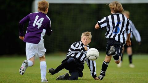 Three boys, playing football. One is on the ground, with a football near to his face, and his eyes closed. Two others have their backs to the camera and are running. Another child is blurred and in the distance.