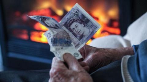 an older person's hands grasp two £20 notes in front of a lit fire