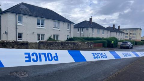 
Blue and white police tape which is upside down in front of a row of cream two-storey buildings. A police officer is visible in a distance.