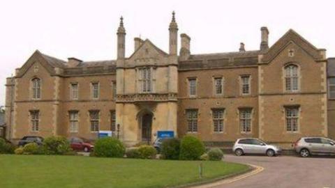 Historic mustard coloured stone building with chimneys and 18 windows and one door. There are cars parked in front of it, some bushes and a grassed area.