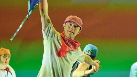 Four dancers wearing t-shirts and peaked caps one is holding aloft a flag, against a background of blurred colours of a rainbow.