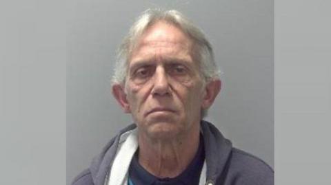 Barry Clarke is standing in front of a grey background. The photo is from the shoulders up, and he is staring into the camera. He has grey hair, and is wearing a grey hoodie and blue polo shirt. 