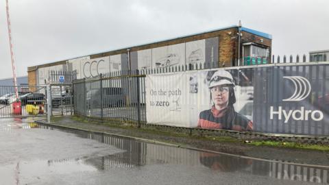 A photo of the Norsk Hydro site in Bedwas, Caerphilly. In the background is a brick building with a metal fence in the foreground. On the fence is a poster with the word Hydro written on it and a picture of a female worker wearing work uniform and a helmet.