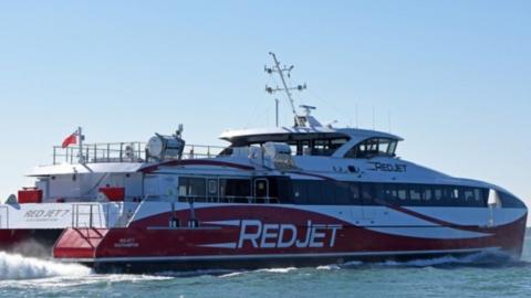 Red and white catamaran travelling on the sea its front it to the right with white spray coming from engines at the rear.