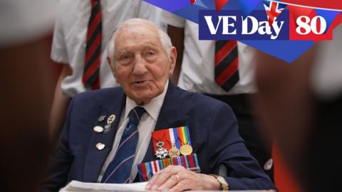 Mervyn Kersh sits surrounded by pupils. He is wearing a navy blue jacket adorned with medals and badges.