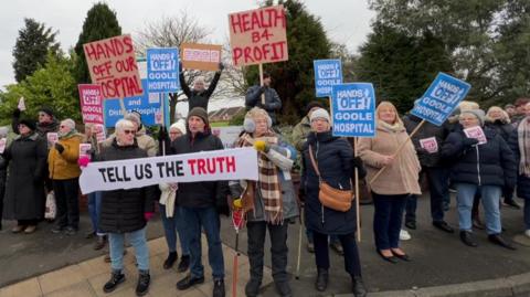 A group of protesters all wearing winter-appropriate clothing. Many are holding protest signs with slogans such as "hands off our Goole hospital". A banner reads "tell us the truth". They are standing on a path in front of a group of tall trees. The sky is overcast. 