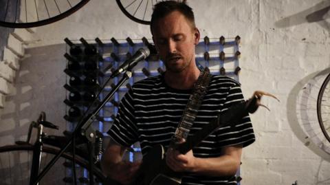 Tom wears a black t-shirt with white stripes. He plays guitar on stage, standing in front of a microphone. A white brick wall is behind him with vintage bikes on display. He has closely-cropped light hair and fair facial stubble.