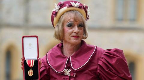 A head and shoulders shot of artist Grayson Perry dressed as his alter ego Claire in a maroon puff-sleeved dress which has yellow border piping. He is wearing a maroon and yellow hat which is adorned with butterflies and bows. He is holding up a red box containing his red and gold Knights Bachelor medal.