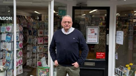 A man standing outside his shop. He is wearing a navy blue jumper over a shirt and glasses. He has his hands tucked into his front pockets and he is looking into the camera. There are greetings cards in the shop window.