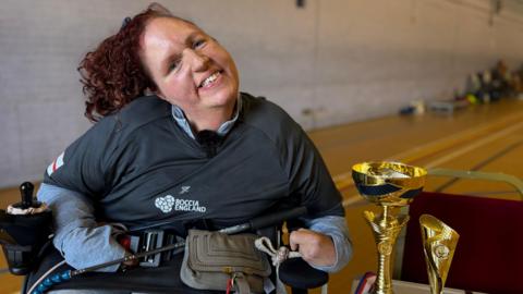 Georgina, who has long, dark red, curly hair, smiles while sitting in a wheelchair next to her two gold trophies. She is wearing a dark-coloured Boccia England T-shirt. She is pictured in a sports hall.