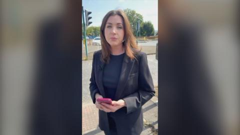 BBC reporter in black jacket by the road.