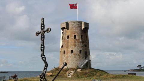 Le Hocq Tower, a round stone building with windows all around it, on a grassy summit with the sea behind it and a large anchor in the foreground 