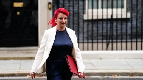Louise Haigh walking out of 10 Downing Street. She has red hair and is wearing a white blazer with a blue dress. She is also carrying a red folder.