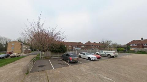 A concrete car park, containing four vehicles, behind a row of modern red brick two-up two-down houses.