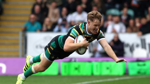 Fin Smith, wearing the Northampton Saints home kit of green, gold and black dives over the try line to score against Leicester