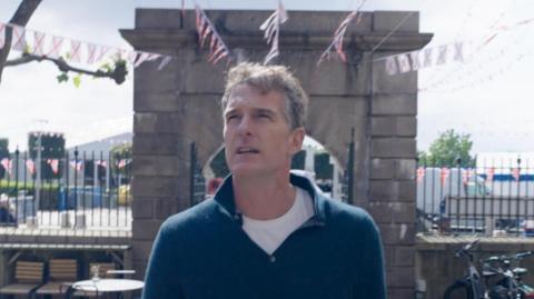 Dan Snow stood in front of a stone archway, looking off to the right. There is bunting behind him and a black metal fence.