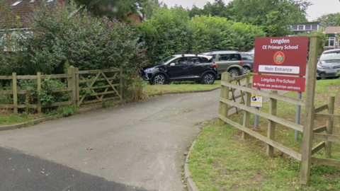 The view towards the entrance to a school, with open wooden fencing on both sides of the road, and a large sign on the right with the words Longden CE Primary School