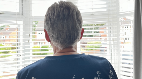 Sue is looking out of her window at home with her back to the camera. She is wearing a blue patterned top and has short grey hair.