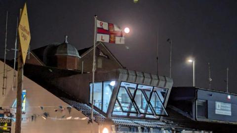 Poole RNLI lifeboat station at night. The red and white RNLI flag is flying on the flagpole and the moon is shining in the sky.
