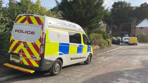 Police van parked half up on a pavement with a smaller van further down to road homes can be seen on the left side